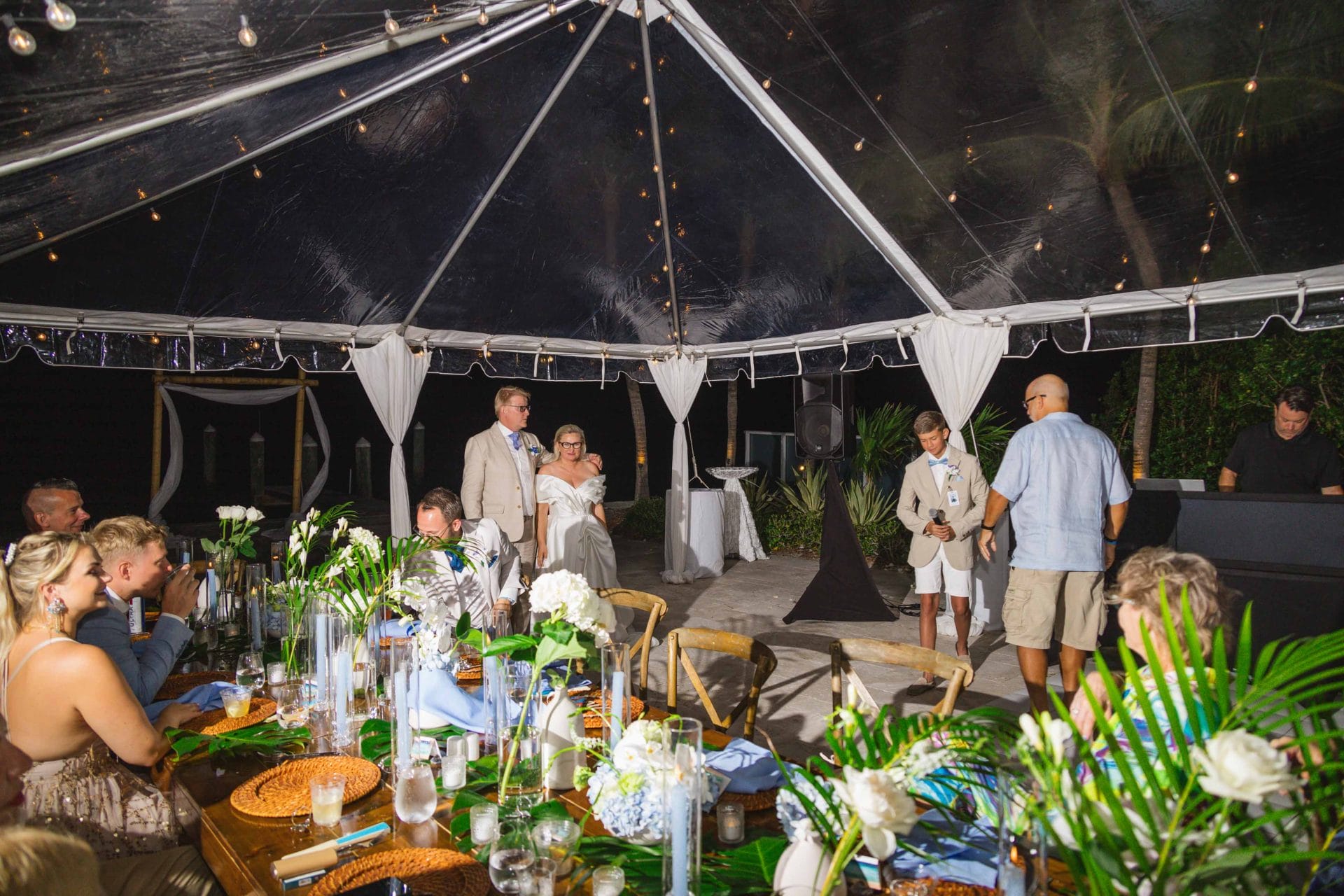 2024-10-29 SOcial media_-1064 A wedding reception under a clear tent at night, with guests seated at tables decorated with flowers and foliage. A couple stands at the front beside a young boy and a speaker.